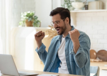 Happy guy celebrate great news opportunity got read by e-mail. Excited man sit in kitchen look at laptop screen with clenched fists feels incredible amazed received good notice. Sale, discount concept