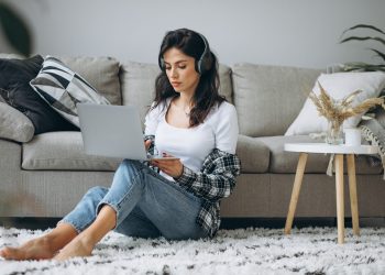 Young beautiful woman sitting home working on laptop in earphones