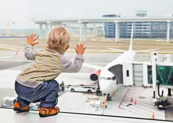 Little baby boy waiting boarding to flight in airport transit hall and looking through the window at airplane near departure gate. Active family lifestyle, travel by air with child on summer vacation.