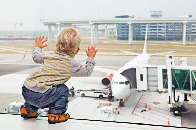 Little baby boy waiting boarding to flight in airport transit hall and looking through the window at airplane near departure gate. Active family lifestyle, travel by air with child on summer vacation.