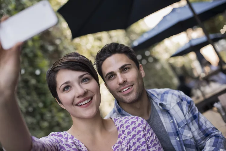 A couple seated at an outdoor city cafe, taking a selfy with a smart phone.