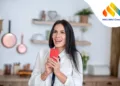 At home.Young woman in her kitechen, holding red smartphone, smiling excitedly