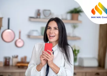 At home.Young woman in her kitechen, holding red smartphone, smiling excitedly