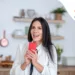 At home.Young woman in her kitechen, holding red smartphone, smiling excitedly