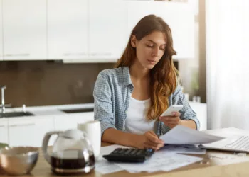 Candid shot of beautiful concentrated young Caucasian woman typing text message on mobile phone while calculating bills in kitchen, sitting at table with laptop computer, papers and calculator