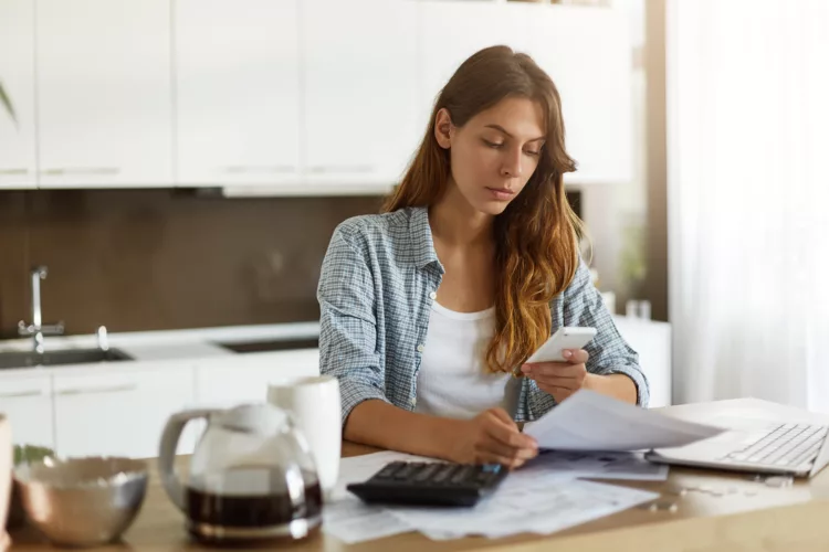 Candid shot of beautiful concentrated young Caucasian woman typing text message on mobile phone while calculating bills in kitchen, sitting at table with laptop computer, papers and calculator