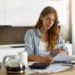 Candid shot of beautiful concentrated young Caucasian woman typing text message on mobile phone while calculating bills in kitchen, sitting at table with laptop computer, papers and calculator