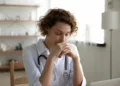 Close up thoughtful woman doctor with closed eyes sitting at work desk in hospital, meditating at workplace, female therapist physician gp wearing white coat uniform praying, asking help