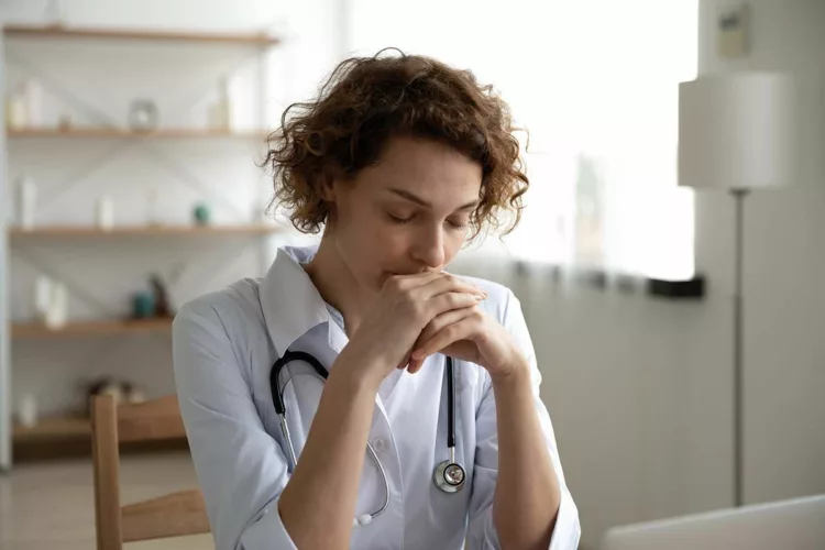 Close up thoughtful woman doctor with closed eyes sitting at work desk in hospital, meditating at workplace, female therapist physician gp wearing white coat uniform praying, asking help