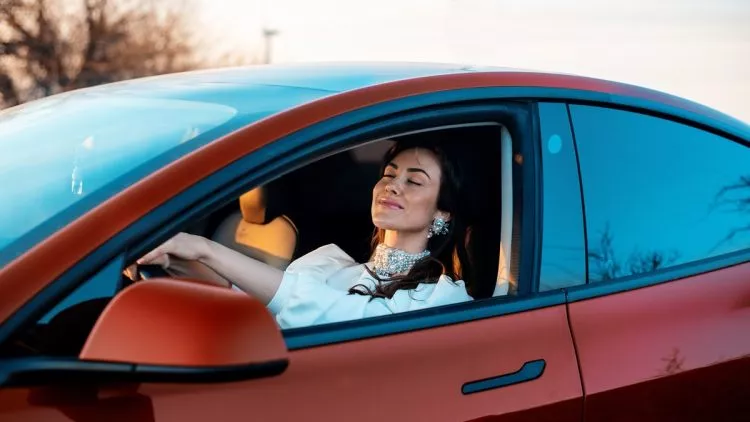 Close view of a woman smiling with closed eyes while sitting in a red parked car at sunset